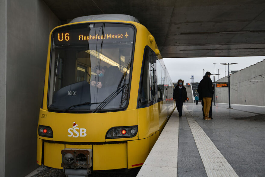 Eine Straßenbahn der SSB steht an der Haltestelle Flughafen/messe, während einige Menschen auf dem Bahnsteig stehen.